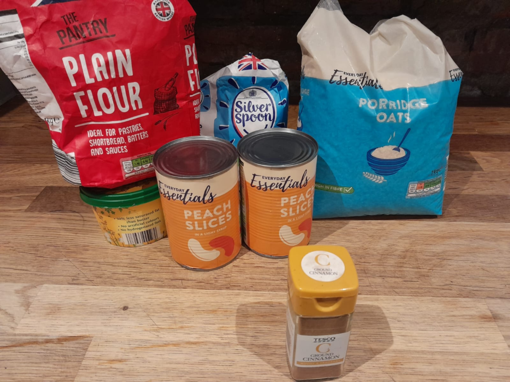 A photo of the ingredients needed to make peach crumble; tinned peaches, plain flour, sugar, butter, porridge oats and cinnamon